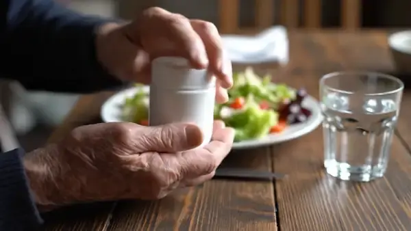 A man's hands tipping a softgel and two capsules from a white jar beside a plate of salad and a glass of water