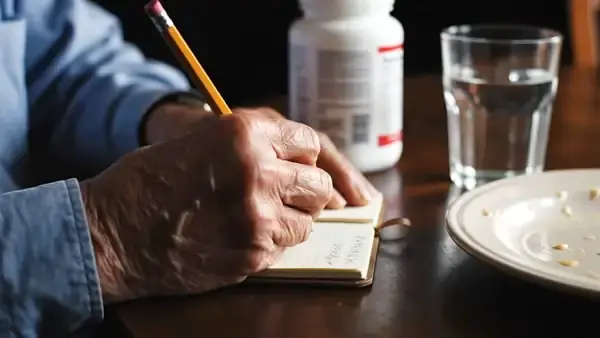 A man's hands writing a line in a small notebook beside a white supplement jar, a glass of water and a lunch plate